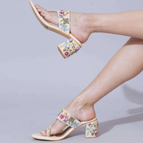 A pair of floral embroidered block heel sandals in ivory color with a 2-inch heel, displayed on a person's feet against a grey background.