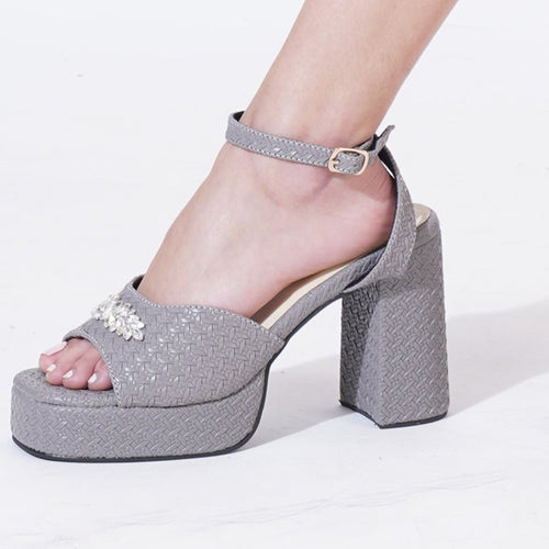 A pair of grey platform heel sandals with a 4-inch block heel, silver brooch embellishments, and ankle straps. The sandals are displayed on a person's feet, indicating a secure fit and a textured woven design.