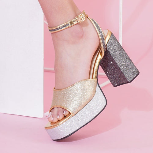 A pair of golden heels with silver accents and a platform heel design, modeled against a pink background.