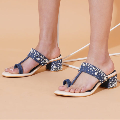 A pair of denim pearl embellished block heels with toe-ring and T-strap detailing, presented against an orange backdrop.